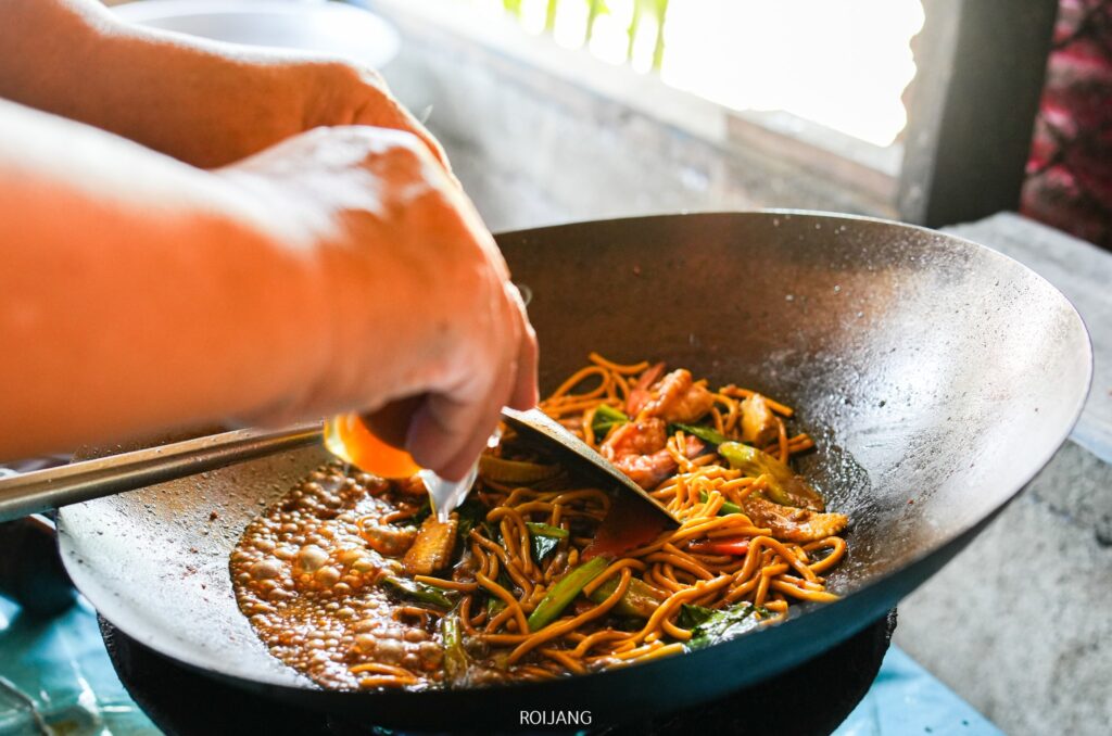 บุคคลหนึ่งทำก๋วยเตี๋ยวผัดผักและเนื้อสัตว์ในกระทะอย่างชำนาญ โดยใช้คีมคีบส่วนผสมที่อร่อยเข้าด้วยกันพร้อมราดซอสที่ชวนให้นึกถึงนับดาวเย็นตาโฟ กลิ่นหอมอบอวลไปทั่วครัวพร้อมกลิ่นอายความเป็นไทย.