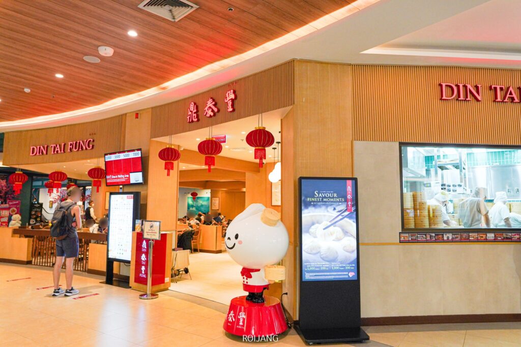 ทางเข้าร้าน Din Tai Fung ภูเก็ต มีป้ายและรูปปั้นตัวละครแปลกตา ภายในร้าน ลูกค้าจะได้ลิ้มรสอาหารในขณะที่เชฟกำลังปรุงอาหารรสเลิศในครัวกระจก โคมแดงที่สาดแสงอบอุ่นส่องลงมายังสวรรค์แห่งอาหารแห่งนี้.