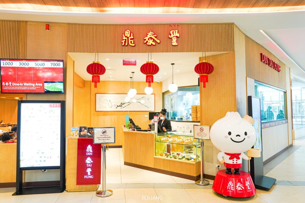 ร้านอาหาร Din Tai Fung ภูเก็ต โดดเด่นด้วยป้ายสีแดงสดและโคมไฟ มีมาสคอตเกี๊ยวแสนน่ารักคอยต้อนรับแขกที่ทางเข้า ส่วนเมนูดิจิทัลทางด้านซ้ายมือจะแนะนำเมนูแสนอร่อยภายในร้าน.