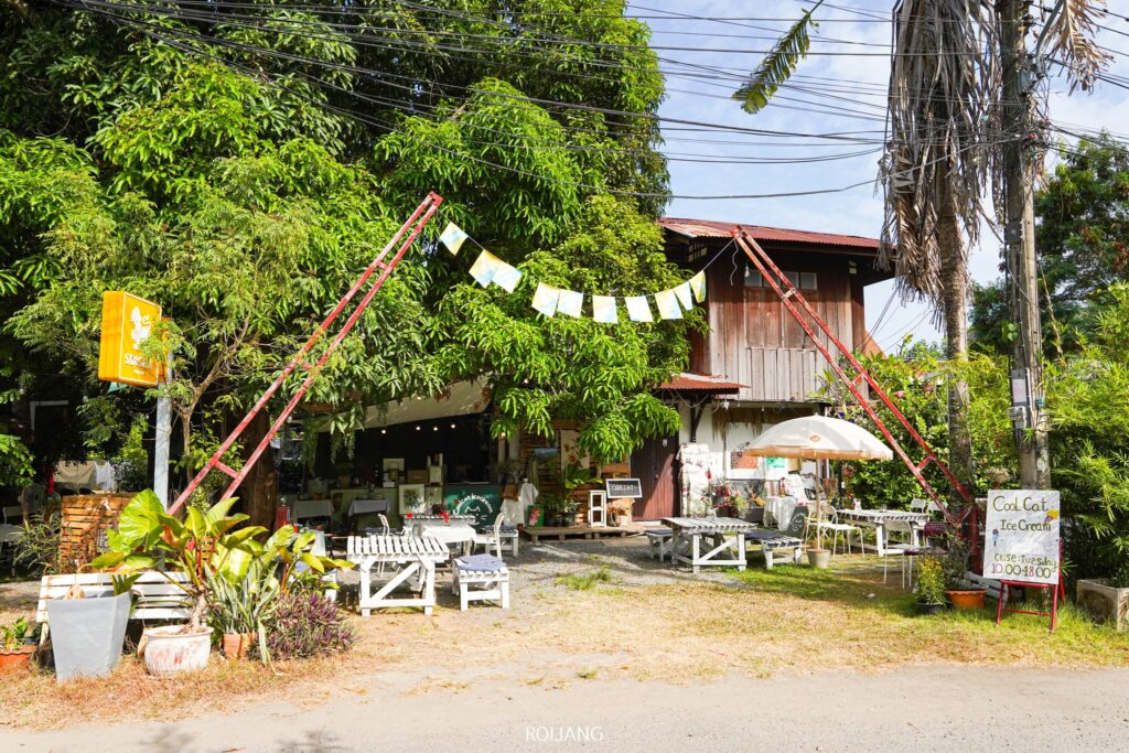 คาเฟ่กลางแจ้งที่มีที่นั่งไม้ใต้ร่มไม้ โครงสร้างสามเหลี่ยมสีแดงตั้งตระหง่านอยู่บริเวณทางเข้า เชื้อเชิญให้คุณเข้าไปลิ้มรสอาหารอันแสนอร่อย ป้ายสีเหลืองและกระดานเมนูแสดงให้เห็นเมนูสุดน่ารับประทานของร้าน Coolcat Icecream.