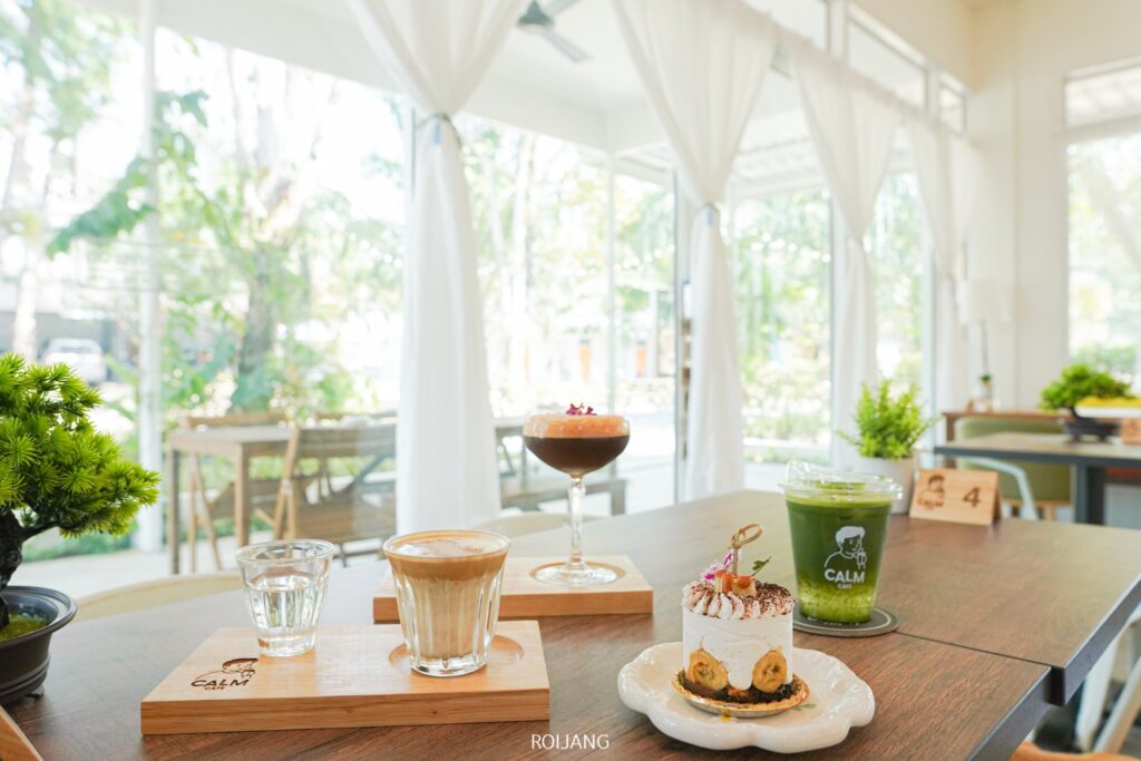 ในบรรยากาศร้านกาแฟอันแสนอบอุ่น "นับดาวเย็นตาโฟ" ประดับประดาบนโต๊ะพร้อมเครื่องดื่มสองแก้วและเค้กหนึ่งชิ้นบนไม้ขัดเงา หน้าต่างบานใหญ่พร้อมผ้าม่านโปร่งเผยให้เห็นความเขียวขจีของต้นไม้ภายนอก สร้างสรรค์การผสมผสานที่ลงตัวระหว่างรสชาติและทิวทัศน์.