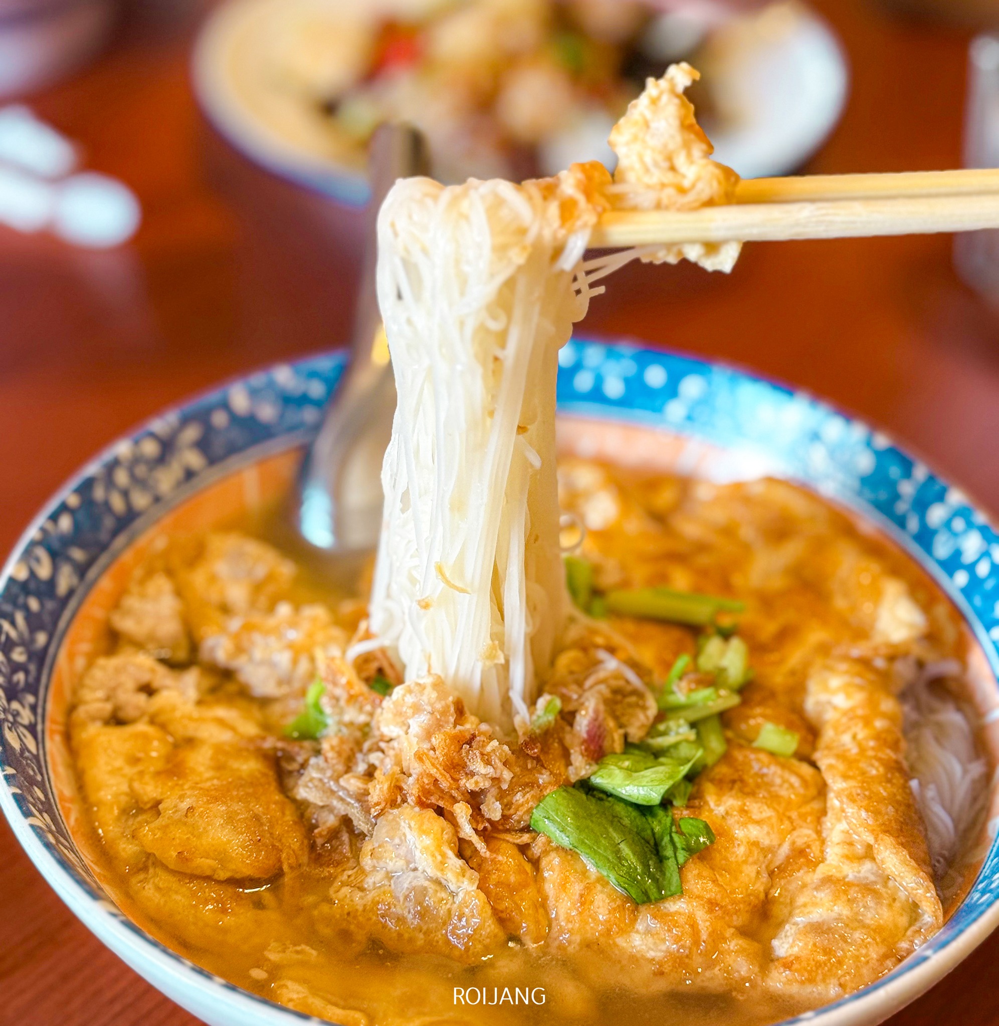 ภาพระยะใกล้ของชามก๋วยเตี๋ยว YiLin ที่มีตะเกียบคีบก๋วยเตี๋ยว ซุปที่เสิร์ฟในชามลายสีน้ำเงินเต็มไปด้วยผัก ไข่ และผักกรุบกรอบ มอบประสบการณ์ที่น่ารื่นรมย์.