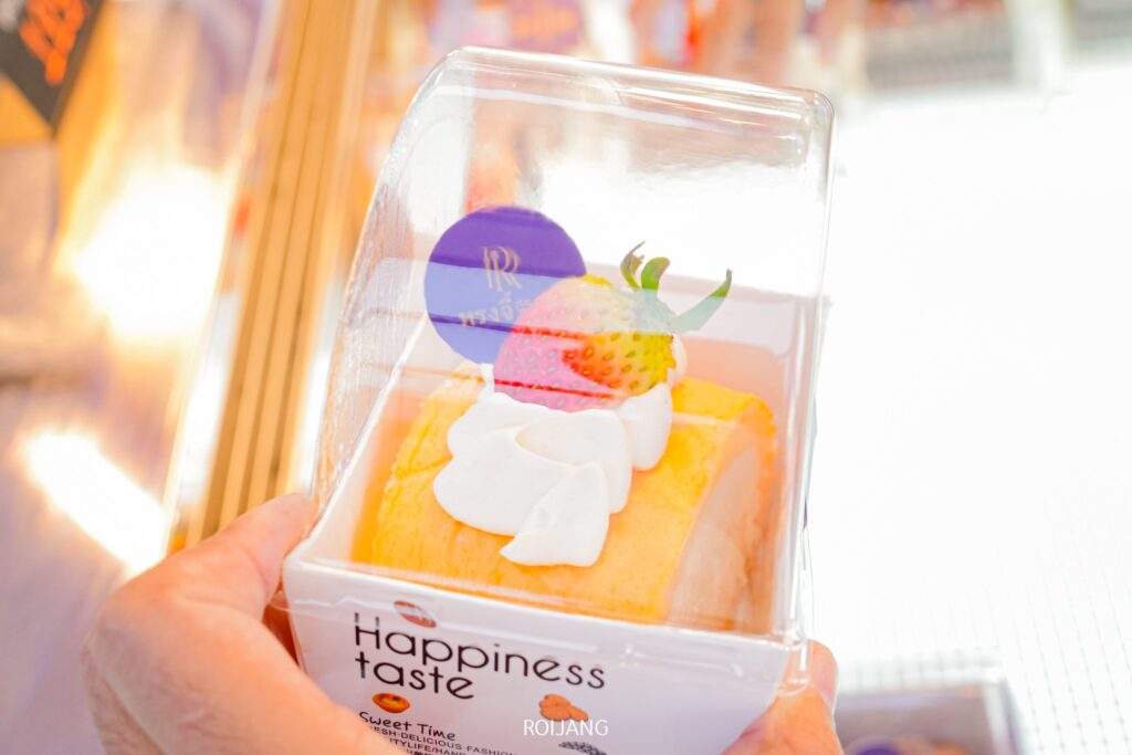 คนถือขนมบรรจุหีบห่อที่ราดด้วยวิปครีมและสตรอว์เบอร์รี บนฉลากเขียนว่า “Happiness taste” เป็นผลงานสร้างสรรค์อันน่ารื่นรมย์จาก RONGJI Bakery ที่ผสมผสานความหวานและความสุขในทุกคำ.