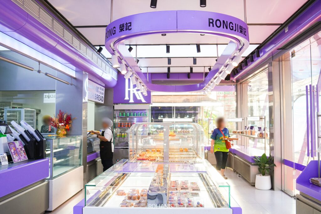 ร้านเบเกอรี่ RONGJI โดดเด่นด้วยกลิ่นอายสีม่วงอันเป็นเอกลักษณ์ โดยมีขนมอบและเค้กวางเรียงรายอยู่ตรงกลางร้าน ในร้านมีลูกค้าคนหนึ่งยืนอยู่ที่เคาน์เตอร์ ขณะที่อีกคนหนึ่งเดินผ่านไปพร้อมกับถาดที่อบอวลไปด้วยกลิ่นหอมเย้ายวนของขนมอบสดใหม่.