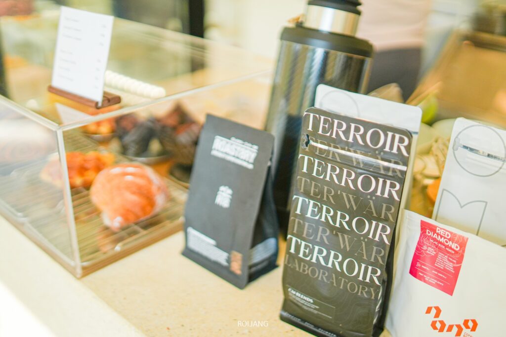 ถุงกาแฟที่มีฉลากว่า “Terroir” และ “Red Diamond” ถูกจัดแสดงไว้อย่างมีศิลปะบนเคาน์เตอร์ที่ Candlery House Cafe ใกล้กับแผนกขนมอบที่น่ารับประทาน พร้อมด้วยกระติกน้ำร้อนดีไซน์เก๋.