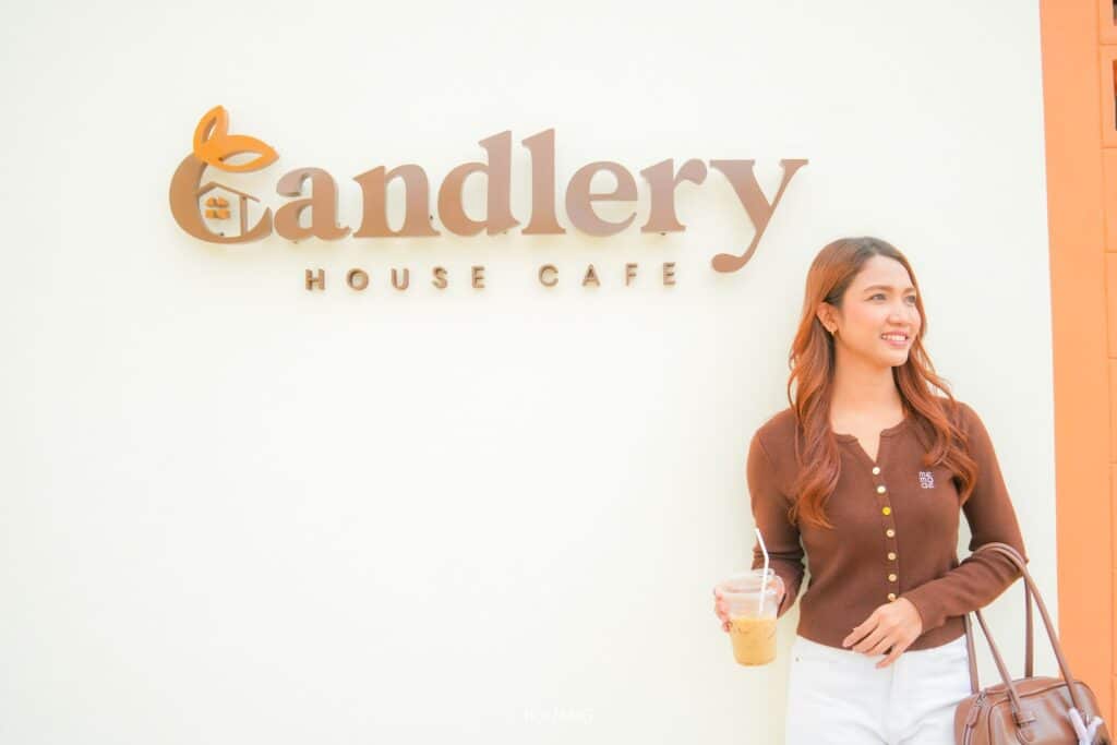 ชายคนหนึ่งถือถ้วยกาแฟที่กำลังร้อนอยู่โพสท่าอย่างร่าเริงต่อหน้าป้าย "Candlery House Cafe" ที่น่าเชิญชวนซึ่งติดอยู่บนผนัง.