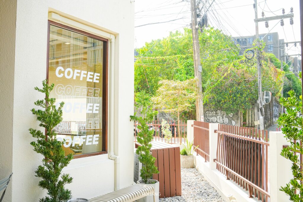 ด้านนอกของร้าน The Candlery House Cafe มีหน้าต่างที่ประดับด้วยคำว่า "COFFEE" ซ้ำๆ กัน ด้านนอกมีกระถางต้นไม้และม้านั่งลายทางที่ชวนให้ผ่อนคลายท่ามกลางทัศนียภาพของต้นไม้เขียวขจีและสายไฟฟ้าที่อยู่ไกลออกไป.