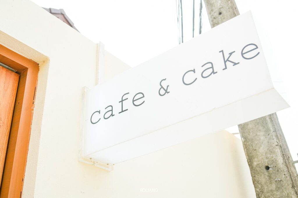 ป้ายสีขาวเขียนว่า “Candlery House Cafe & Cake” ติดไว้อย่างหรูหราบนผนังสีอ่อนข้างประตูไม้ที่สวยงาม.