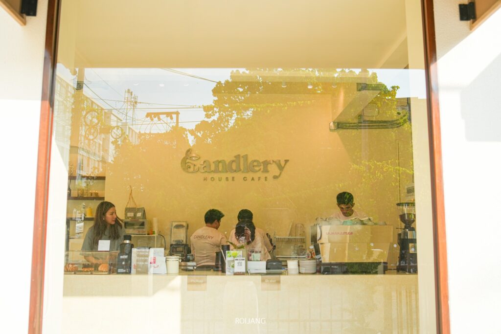 มุมมองภายนอกของร้าน Candlery House Cafe เผยให้เห็นพนักงาน 4 คนที่กำลังนั่งทำงานอย่างขะมักเขม้นอยู่หลังเคาน์เตอร์ ชงกาแฟและเตรียมออเดอร์อย่างชำนาญ โดยมีชื่อร้านกาแฟแสดงอยู่บนผนังด้านหลังอย่างสวยงาม.