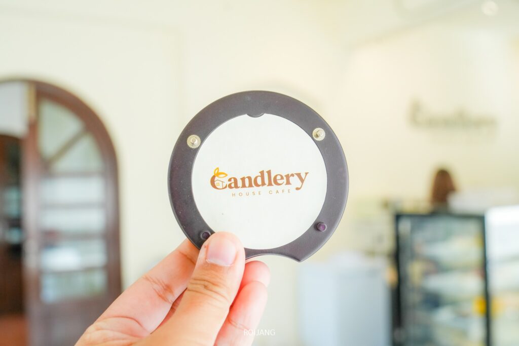 มือถือเพจเจอร์ทรงกลมที่ประดับโลโก้ "Candlery House Cafe" บนฉากหลังร้านกาแฟที่เบลอเล็กน้อย.