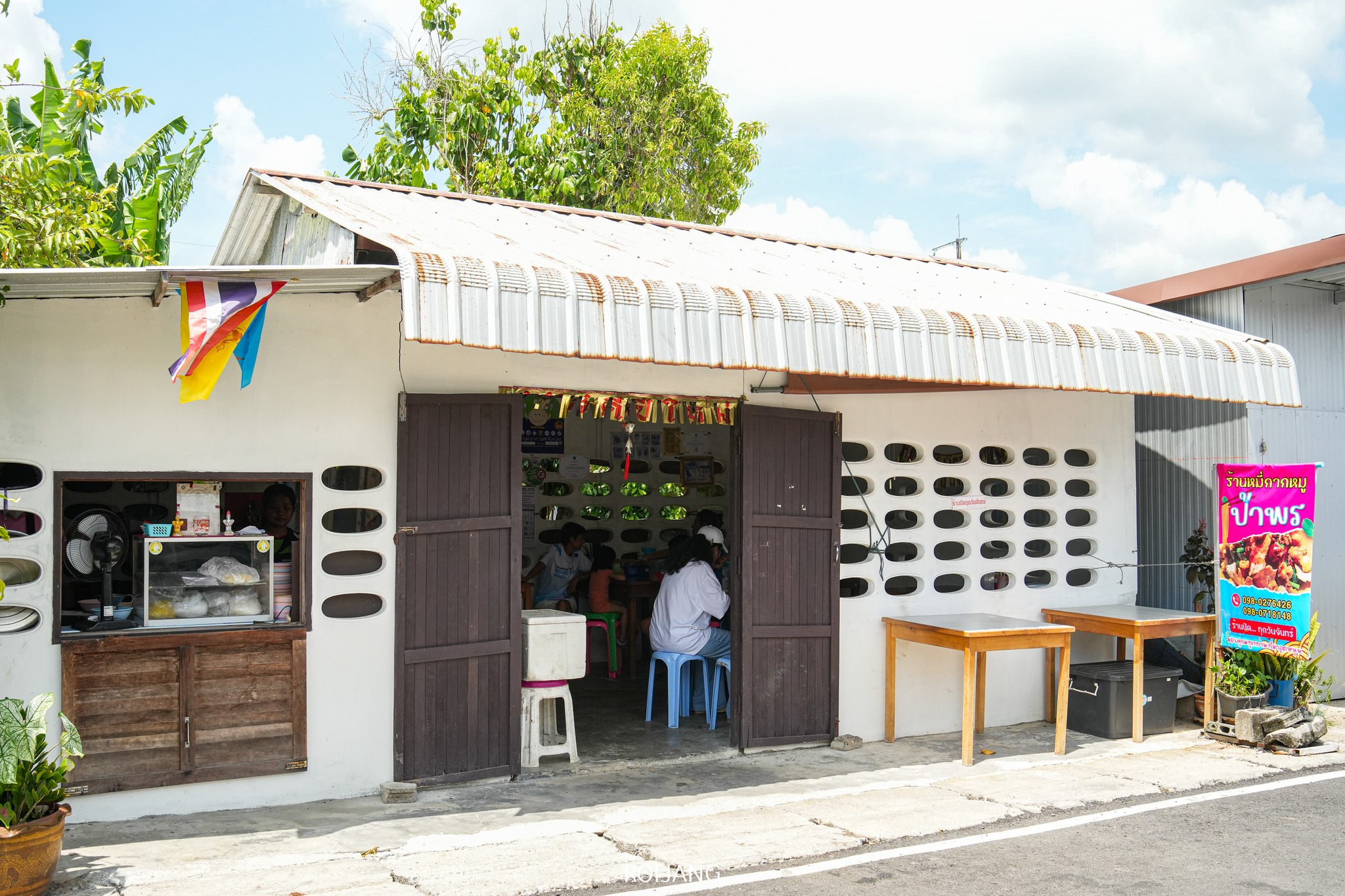 ร้านหมี่กากหมูป้าพร เป็นร้านอาหารเล็กๆ ริมถนนสีขาว มีประตูไม้และหลังคามุงด้วยแผ่นไม้เปิดโล่ง ภายในร้านมีลูกค้านั่งรับประทานอาหารอยู่หลายคน ป้ายสีสันสดใสและธงสีสันสดใสต้อนรับแขกที่ทางเข้า.