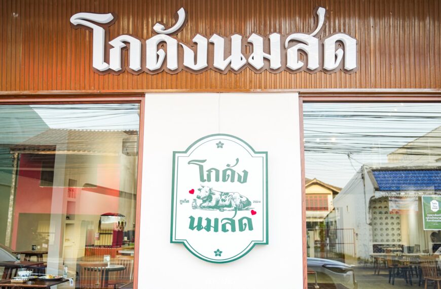 ภายนอกของร้านอาหารมีด้านหน้าเป็นไม้ที่สวยงาม ตกแต่งด้วยป้ายอักษรไทยและโลโก้รูปวัวซึ่งชวนให้นึกถึงโกดัง นมสด เงาของอาคารใกล้เคียงส่องประกายในกระจก ช่วยเพิ่มความรู้สึกแบบเมือง.