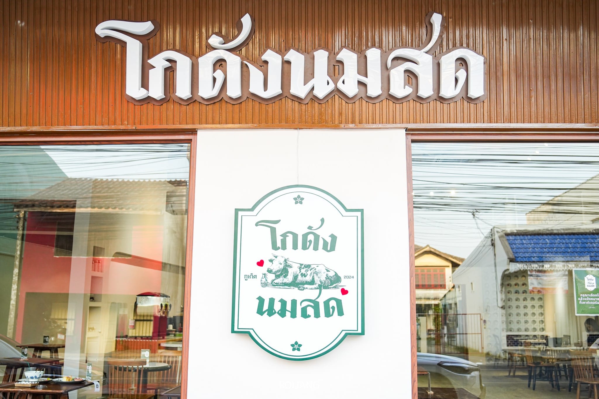 ภายนอกของร้านอาหารมีด้านหน้าเป็นไม้ที่สวยงาม ตกแต่งด้วยป้ายอักษรไทยและโลโก้รูปวัวซึ่งชวนให้นึกถึงโกดัง นมสด เงาของอาคารใกล้เคียงส่องประกายในกระจก ช่วยเพิ่มความรู้สึกแบบเมือง.