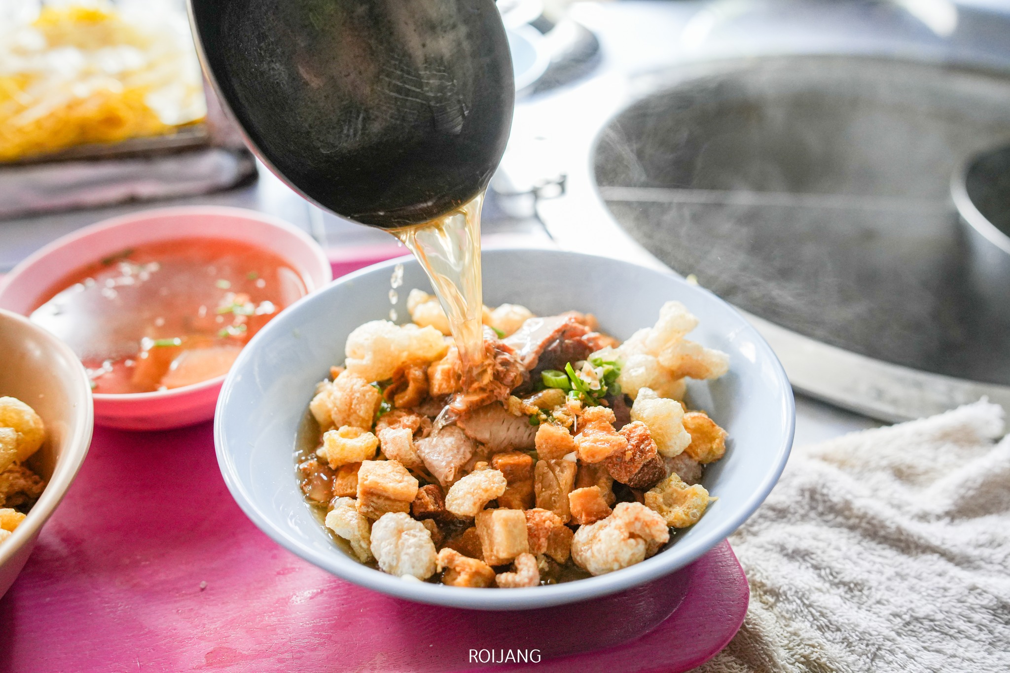 ชามก๋วยเตี๋ยวหมี่กากหมูป้าพร ราดด้วยหมูกรอบ น้ำซุปร้อนๆ ที่ถูกเทจากทัพพี ใกล้ๆ กันมีชามน้ำจิ้มวางอยู่บนถาดสีชมพู พร้อมเสิร์ฟคู่กับอาหารจานอร่อยนี้.