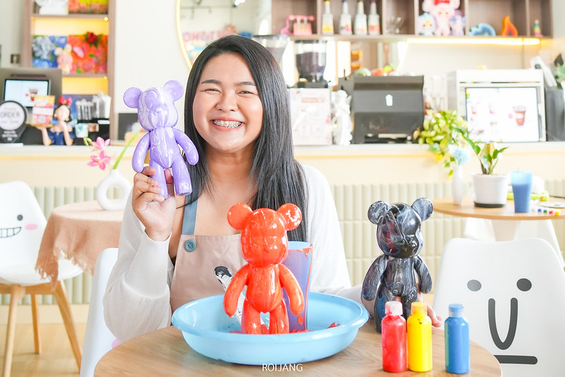 ในร้าน Art & Toy Cafe เมืองพังงา ผู้หญิงคนหนึ่งยิ้มขณะถือตุ๊กตาหมีสีม่วง ล้อมรอบด้วยอุปกรณ์ศิลปะสีสันสดใส และรูปปั้นตุ๊กตาหมีสำเร็จรูปมากมายหลายแบบ.