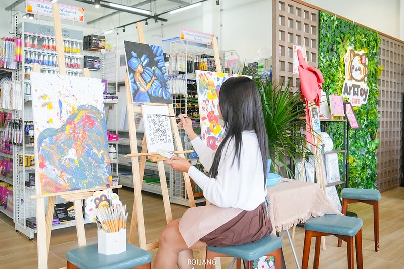 ใน Art & Toy Cafe Phangnga ที่มีชีวิตชีวา ผู้หญิงคนหนึ่งกำลังนั่งที่โต๊ะเล็กๆ ในสตูดิโอศิลปะและกำลังวาดภาพบนผืนผ้าใบ รอบๆ ตัวเธอมีอุปกรณ์ศิลปะและภาพวาดที่ทำเสร็จแล้วมากมาย สร้างสรรค์ฉากหลังที่กระตุ้นความคิดสร้างสรรค์.