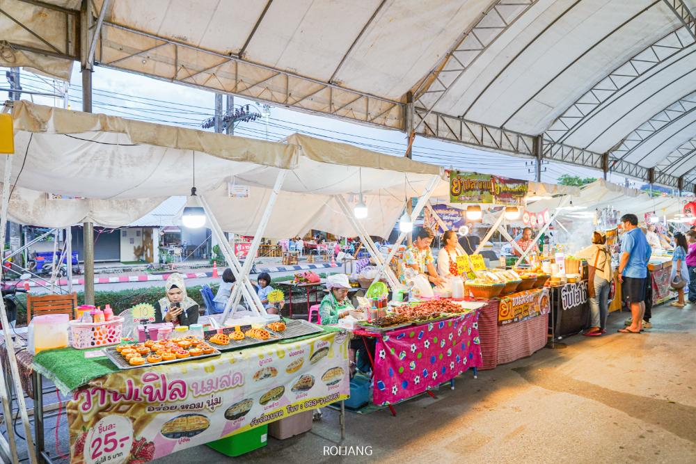 ตลาดกลางแจ้งที่คึกคักเปิดโล่งภายใต้หลังคาสีขาวชวนให้นึกถึงงานบวงสรวงพระนารายณ์ 2568 โดยมีแผงขายของที่นำอาหารและสินค้าหลากหลายมาขาย นักช้อปเดินเตร่ไปตามสินค้าสีสันสดใสเบื้องหลัง.