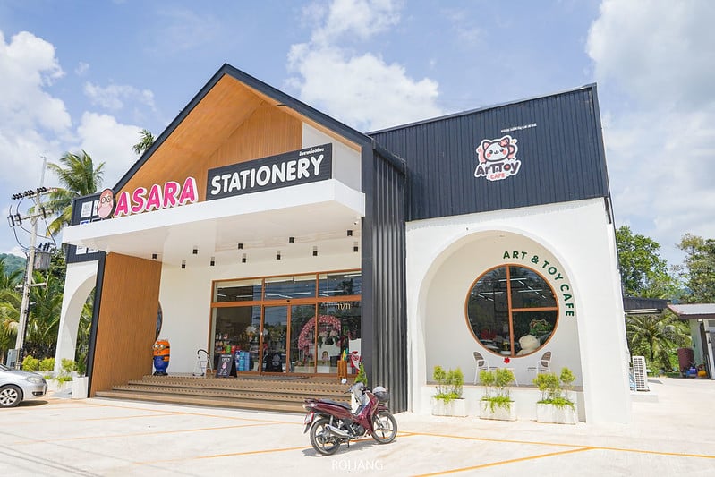 ภายนอกร้าน Asara Stationery and Art & Toy Cafe ที่พังงา ซึ่งเป็นอาคารทันสมัยที่มีหลังคาทรงจั่วและทางเข้าเป็นกระจกขนาดใหญ่ ล้อมรอบด้วยลานจอดรถพร้อมรถจักรยานยนต์ที่จอดอยู่.