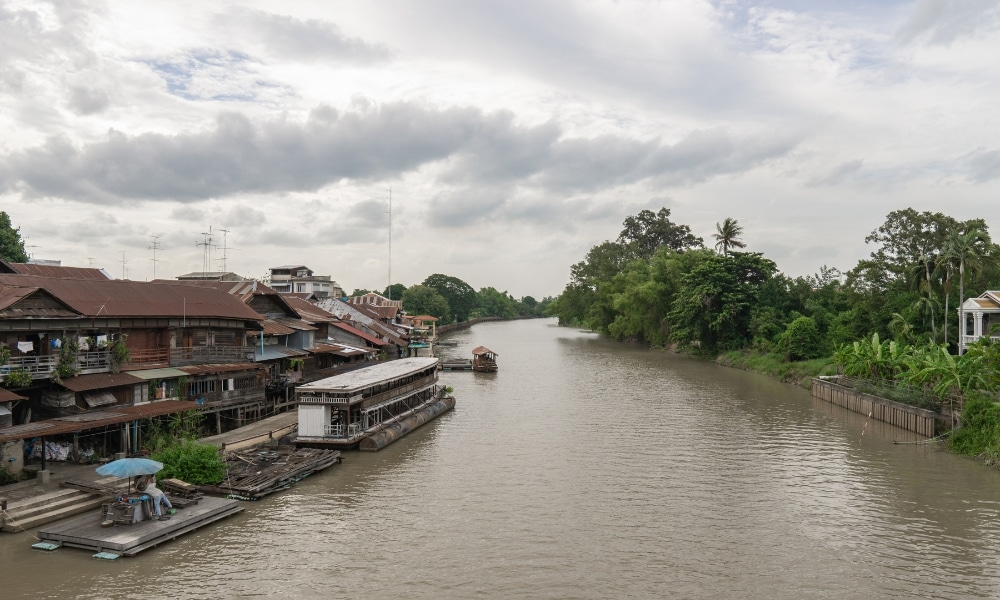 สัมผัสทัศนียภาพริมแม่น้ำอันเงียบสงบของลำน้ำแคบๆ ที่เที่ยวสมุทรสงคราม โดยมีบ้านเรือนอันมีเสน่ห์อยู่ทางซ้ายและต้นไม้เขียวขจีอยู่ทางขวาภายใต้ท้องฟ้าที่มีเมฆครึ้ม โครงสร้างลอยน้ำขนาดเล็กจอดทอดสมออยู่ข้างบ้านเรือน ช่วยเพิ่มสัมผัสอันเงียบสงบให้กับสถานที่นี้.