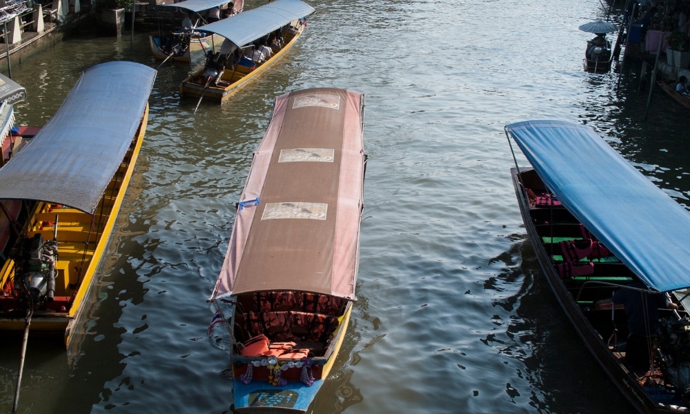 เรือยนต์สี่ลำที่มีหลังคาหลากสีสันแล่นอยู่ใกล้กันบนลำคลองที่ล้อมรอบด้วยอาคารทั้งสองฝั่ง นำเสนอภาพชีวิต สมุทรสงครามที่เที่ยว ประจำวันที่งดงามในที่เที่ยวสมุทรสงคราม.