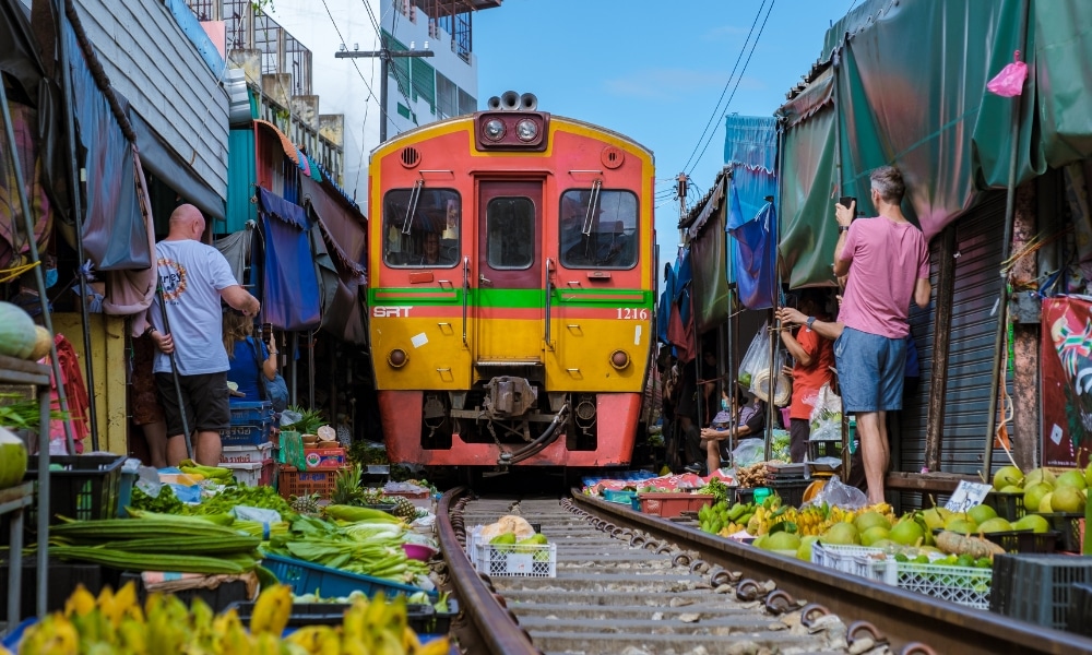 รถไฟแล่นผ่านตลาดแคบๆ ในจังหวัดสมุทรสงคราม โดยมีแผงขายของที่เต็มเปี่ยมไปด้วยผลไม้และผักอยู่สอง ที่เที่ยวสมุทรสงคราม ข้างทาง นักช้อปและพ่อค้าแม่ค้ายืนเบียดเสียดกันใกล้รางรถไฟ ขณะที่กันสาดสีสันสดใสช่วยเพิ่มความมีชีวิตชีวาให้กับสถานที่ท่องเที่ยวที่ไม่เหมือนใครแห่งนี้.