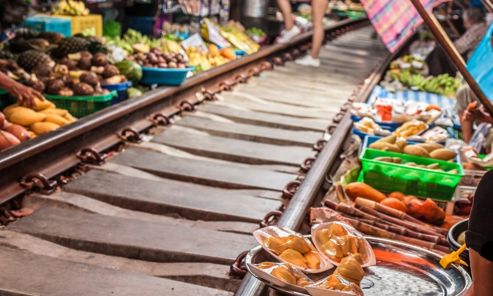 รางรถไฟทอดยาวผ่านตลาดที่คึกคักในที่เที่ยวสมุทรสงคราม โดยมีผักและ ที่เที่ยวสมุทรสงคราม ผลไม้สีสันสดใสขึ้นทั้งสองด้าน ร่มหลากสีสันให้ร่มเงาเมื่อผู้คนกลมกลืนไปกับฉากหลังที่มีชีวิตชีวา.