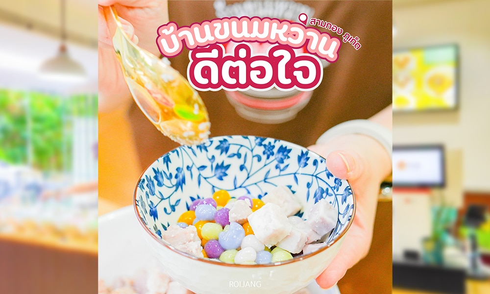 ที่บ้านขนมหวาน มีคนเสิร์ฟของหวานหลากสีสันที่ตกแต่งด้วยท็อปปิ้งทรงกลมเล็กๆ ในชามสีน้ำเงินและสีขาว บรรยากาศภายในอาคารที่พร่ามัวช่วยเพิ่มเสน่ห์ ขณะที่ป้ายบอกเป็นนัยถึงจุดที่น่ารื่นรมย์ในภูเก็ตซึ่งดีต่อใจอย่างแท้จริง.