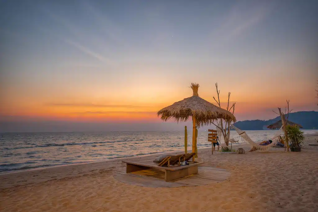ฉากชายหาดอันเงียบสงบยาม ที่พักหาดเจ้าหลาว พระอาทิตย์ตกดินบนหาดเจ้าหลาวติดทะเล มีร่มมุงจาก พื้นที่เลานจ์ไม้ และเปลญวน เฉดสีอบอุ่นแต่งแต้มท้องฟ้าขณะที่ทะเลอันเงียบสงบทอดยาวไปจนถึงขอบฟ้า.