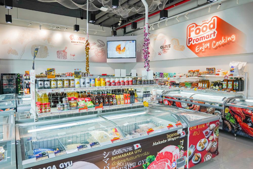 ทางเดินในซูเปอร์มาร์เก็ตที่สว่างไสวของ Food Promart Phuket จัดแสดงผลิตภัณฑ์อาหารต่างๆ รวมถึงอาหารแช่แข็ง ขนมขบเคี้ยว และอุปกรณ์ประกอบอาหาร ป้ายและของตกแต่งสีสันสดใสช่วยเพิ่มสีสันให้กับพื้นที่ที่จัดอย่างเป็นระเบียบ.