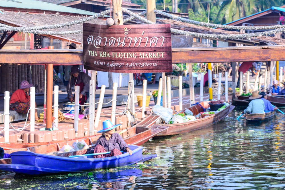 เรือพร้อมสินค้าลอยอยู่ใต้ป้ายไม้ที่ตลาดน้ำท่าคาในประเทศไทย จุดที่มี ที่เที่ยวสมุทรสงคราม  ที่เที่ยวสมุทรสงคราม เสน่ห์ที่เที่ยวสมุทรสงคราม ผู้คนสวมหมวกล่องไปตามทางน้ำท่ามกลางสภาพแวดล้อมเขตร้อน ช่วยเพิ่มประสบการณ์ท้องถิ่นที่มีชีวิตชีวา.