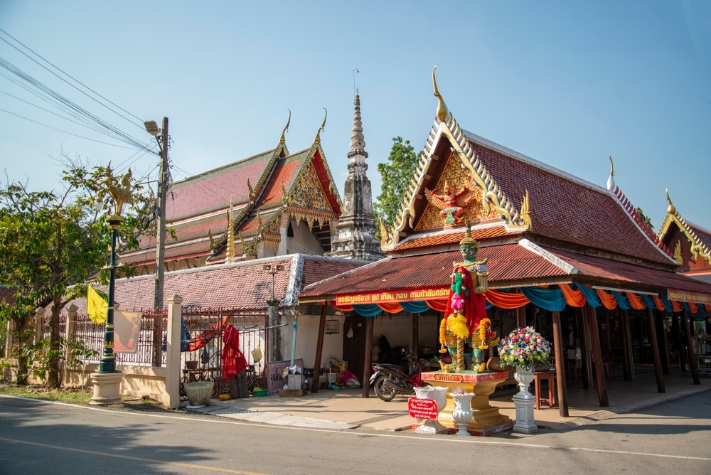 วัดไทยแบบดั้งเดิมที่เที่ยว ที่เที่ยวสมุทรสงคราม สมุทรสงครามมีหลังคาหรูหรา ยอดแหลมตรงกลาง และรูปปั้นเล็กๆ ที่ประดับด้วยดอกไม้ที่ทางเข้า บริเวณนี้อาบไปด้วยแสงแดดภายใต้ท้องฟ้าแจ่มใส.