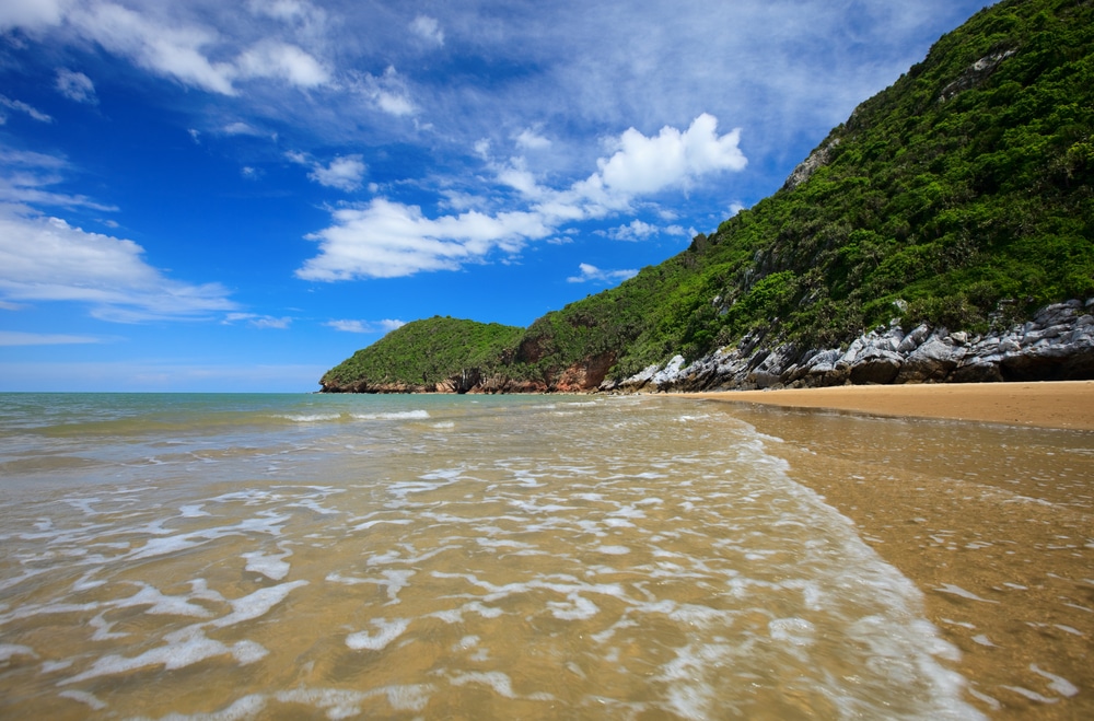 หาดทรายที่มีคลื่นอ่อนโยนบรรจบชายฝั่ง ล้อมรอบด้วยเนินเขาสีเขียว ที่เที่ยวหัวหิน ชอุ่มภายใต้ท้องฟ้าสีครามที่มีเมฆบางส่วน มอบการหลบหนีที่สมบูรณ์แบบเมื่อสำรวจที่เที่ยวหัวหิน.