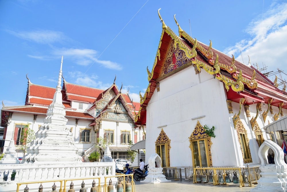 วัดไทยที่มีหลังคาสีแดงและสีทองอันวิจิตรงดงาม การแกะสลักที่สลับซับ สมุทรสงครามที่เที่ยว ซ้อน และเจดีย์สีขาวขนาดใหญ่ที่อยู่เบื้องหน้า ตั้งตระหง่านอย่างสง่างามภายใต้ท้องฟ้าสีฟ้าใสของกองทัพเรือ.