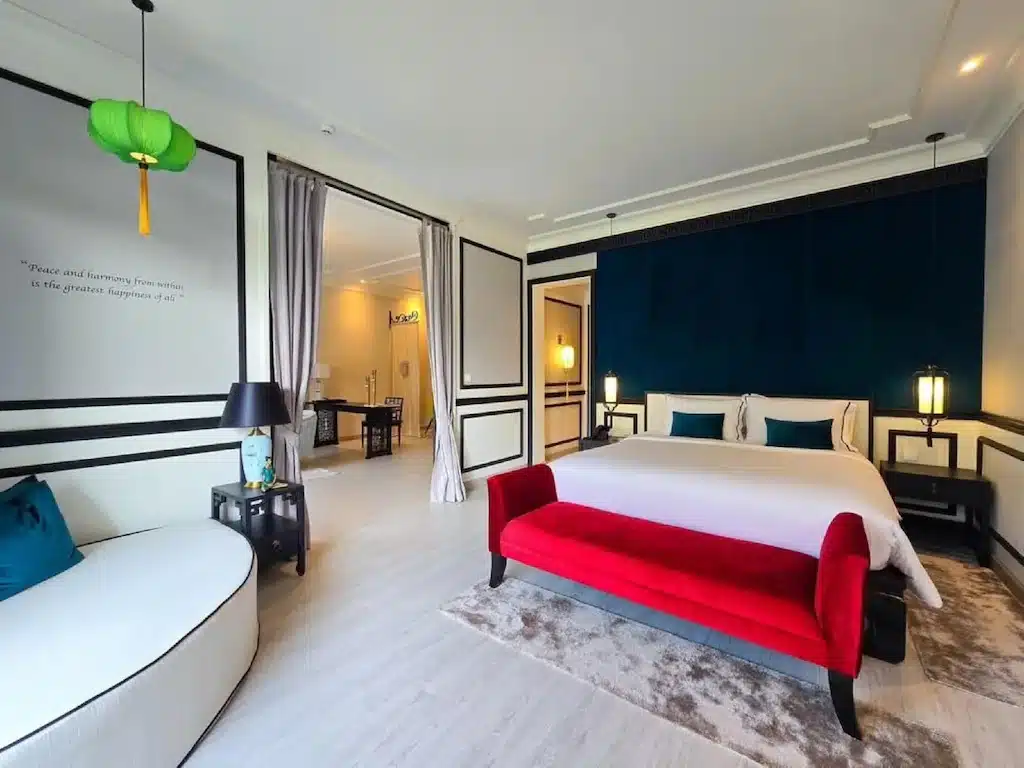 ห้องพักในโรงแรมทันสมัยมีเตียง ที่เที่ยวแก่งคอย ขนาดใหญ่พร้อมผ้าปูเตียงสีขาว ม้านั่งสีแดง โซฟา โคมไฟสองดวง และวิวพื้นที่รับประทานอาหารที่อยู่ติดกัน เหมาะสำหรับการพักผ่อนหลังจากเที่ยวชมที่เที่ยวแก่งคอย.