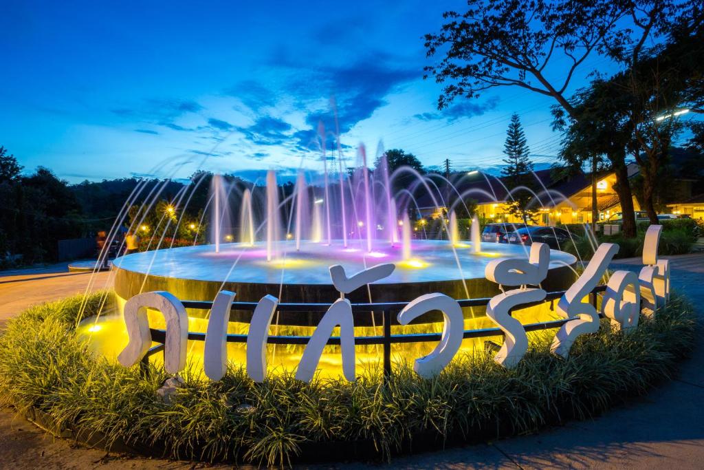 น้ำพุทรงกลมพร้อมไฟหลากสีสันและสายน้ำล้อมรอบด้วยอักษรไทยสีขาว  ที่พักเขาค้อ ต้นไม้และท้องฟ้ายามเย็นสร้างฉากหลังอันเงียบสงบ สะท้อนถึงบรรยากาศที่ผ่อนคลายของหาดเจ้าหลาวติดทะเลในบริเวณใกล้เคียง.