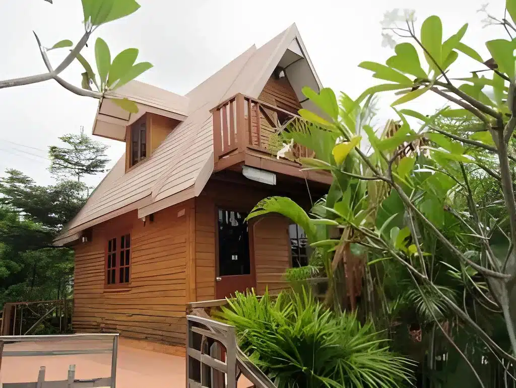 บ้านไม้ที่มีหลังคาและระเบียงสูงชัน  ที่พักเขาค้อราคาถูก ล้อมรอบด้วยต้นไม้สีเขียวและต้นไม้ใต้ท้องฟ้าที่มีเมฆมาก เหมาะสำหรับผู้ที่กำลังมองหาหาดเจ้าหลาวติดทะเลที่โอบล้อมด้วยธรรมชาติ.