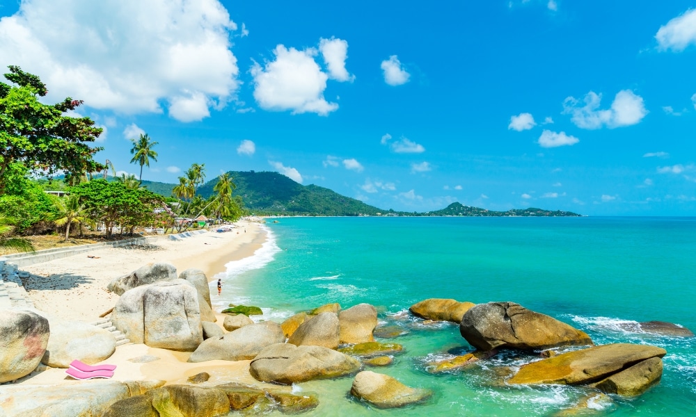 ชายหาดเขตร้อนในที่เที่ยวสมุยที่มีน้ำทะเลสีฟ้าคราม หินขนาดใหญ่ ทรายขาว ต้นปาล์ม และเนินเขาเขียวขจีที่อยู่ห่างไกลภายใต้ท้องฟ้าสีครามและมีเมฆกระจัดกระจาย เหมาะสำหรับการผจญภัยครั้งถัดไปของคุณ ที่เที่ยวเกาะสมุย.