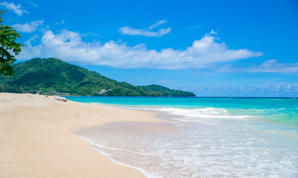 ชายหาดทรายสีฟ้าใส น้ำทะเลใส คลื่นเบาๆ รายล้อมด้วยเนินเขาสีเขียว ท้องฟ้าสีฟ้า มีเมฆเป็นบางส่วน เหมาะสำหรับใครก็ตามที่กำลังมองหา ที่เที่ยวเกาะสมุย และพักผ่อนบนเกาะที่น่าประทับใจ.