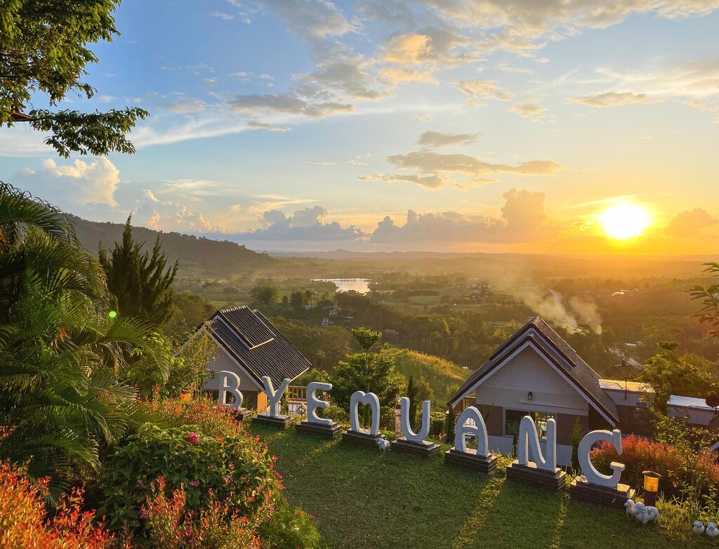 วิวพระอาทิตย์ เขาค้อที่พัก ขึ้นเหนือหุบเขาอันเขียวชอุ่มที่มีบ้านเรือนและตัวอักษร "BYEMUANG" สีขาวขนาดใหญ่อยู่เบื้องหน้า ชวนให้นึกถึงความเงียบสงบที่พบในหาดเจ้าหลาวติดทะเล.