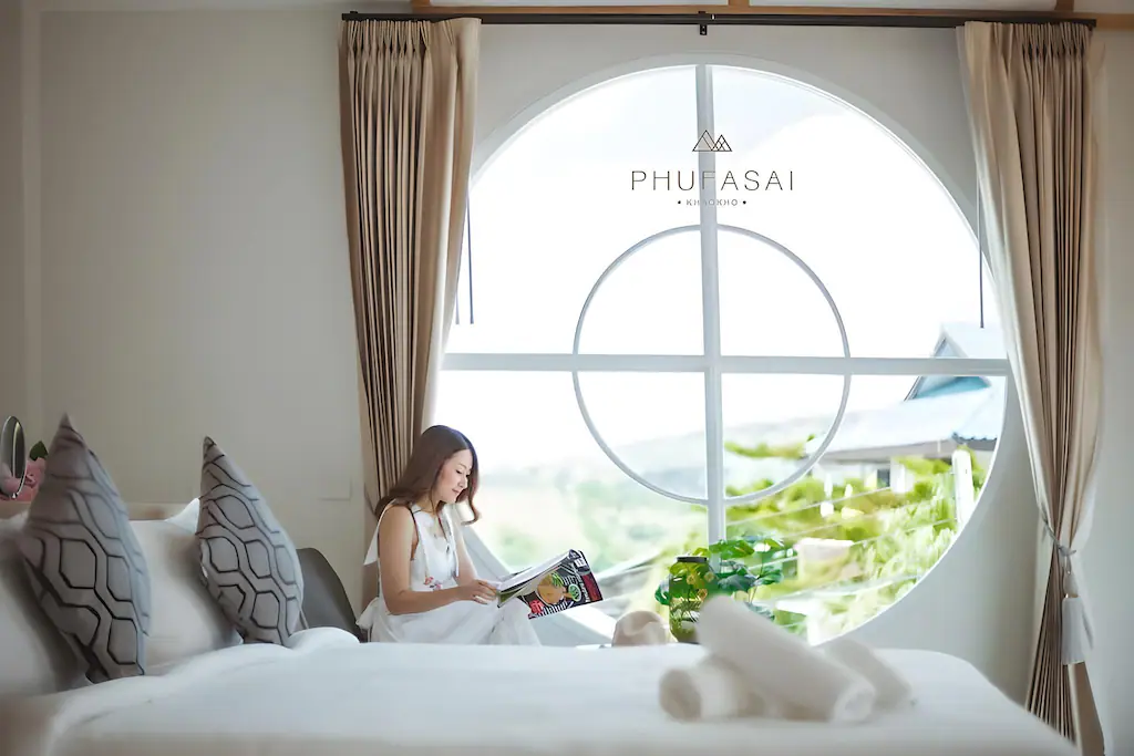 ผู้หญิงคนหนึ่งนั่งอยู่ริมหน้าต่างทรงกลมขนาดใหญ่อ่านนิตยสารในห้องนอนที่ทันสมัยและสว่างสดใสพร้อมการตกแต่งที่เป็นกลางและมี ที่พักเขาค้อ ผ้าเช็ดตัวที่จัดไว้อย่างประณีตบนเตียงที่เจ้าหลาว เหมาะสำหรับการพักผ่อนอย่างผ่อนคลายใกล้ชายหาดเจ้าหลาวที่พัก.