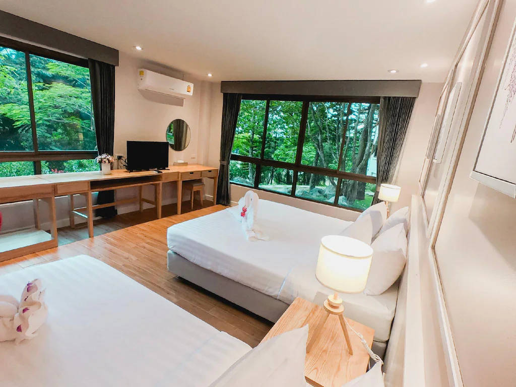 ห้องพักในโรงแรมสว่างสดใสพร้อมเตียง 2 เตียง โต๊ะทำงาน ทีวี และ ที่พักเขาค้อ หน้าต่างบานใหญ่ที่มองเห็นวิวต้นไม้สีเขียว เหมาะสำหรับผู้ที่มองหาหาดเจ้าหลาวติดทะเล โดยมีผ้าเช็ดตัวพับเป็นรูปสัตว์ในแต่ละเตียง.