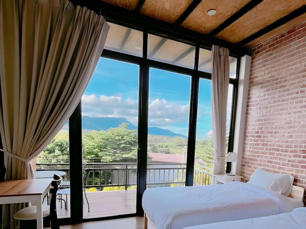 ห้องพักในโรงแรมที่มีเตียงแฝด 2 เตียง ประตูกระจกขนาดใหญ่ ผนังอิฐ และระเบียงที่มองเห็นต้นไม้และภูเขา ที่พักเขาค้อ ไกลๆ ภายใต้ท้องฟ้าสีฟ้า เหมาะสำหรับผู้ที่กำลังมองหาที่พักบนหาดเจ้าหลาวที่มีทิวทัศน์เงียบสงบและสะดวกสบาย.
