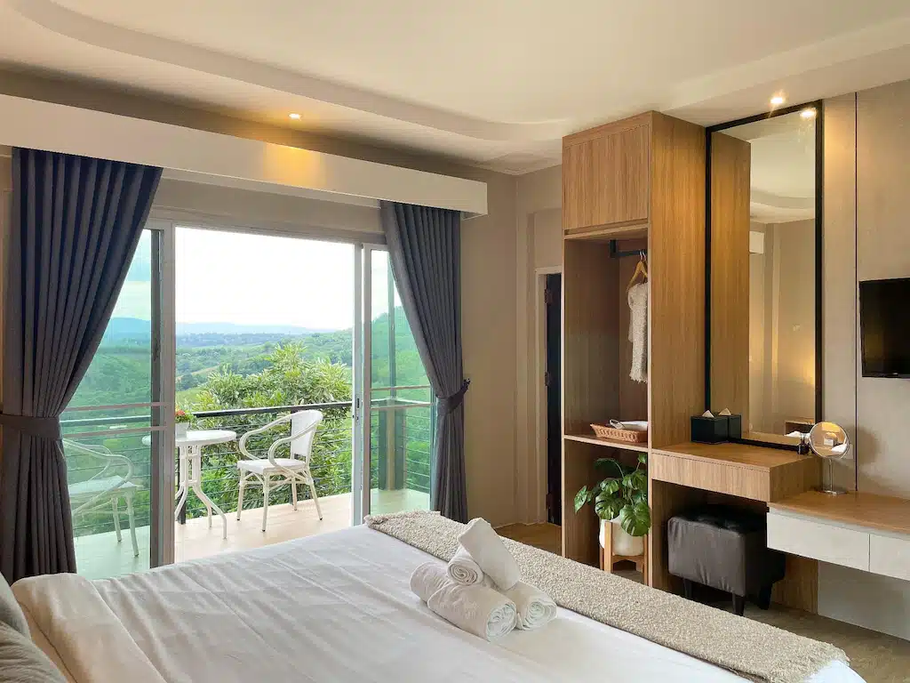 ห้องพักในโรงแรม เขาค้อที่พัก ทันสมัยที่มีเตียง ตู้เสื้อผ้า และโต๊ะทำงานที่ตกแต่งอย่างประณีต พร้อมด้วยประตูกระจกบานเลื่อนที่เปิดออกสู่ระเบียง เหมาะสำหรับการพักผ่อนที่หาดเจ้าหลาวติดทะเล มองเห็นเนินเขาเขียวขจี และบรรยากาศชายหาดอันเงียบสงบ.