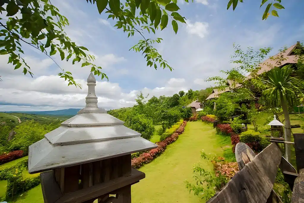 สวนภูมิทัศน์ที่มีหญ้าสีเขียว พุ่มไม้ตัดแต่ง และโครงสร้างหลังคาโลหะยินดี ที่พักเขาค้อ ต้อนรับคุณที่หาดเจ้าหลาวติดทะเล ตั้งอยู่บนเนินเขาใต้ท้องฟ้าที่มีเมฆบางส่วน.