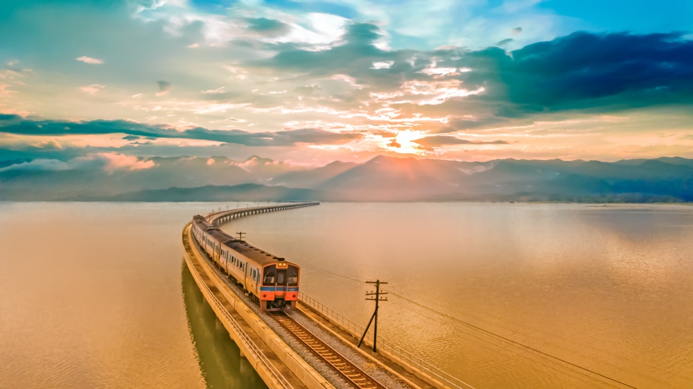 รถไฟโดยสารเดินทางบนสะพานยาวเหนือน้ำยามพระอาทิตย์ตกดิน โดยมีภูเขาและเมฆเป็นฉากหลัง ทิวทัศน์ ที่เที่ยวแก่งคอย อันงดงามเหมาะสำหรับผู้ที่กำลังมองหาที่เที่ยวแก่งคอย หรือสำรวจที่เที่ยวชะอมแก่งคอย.