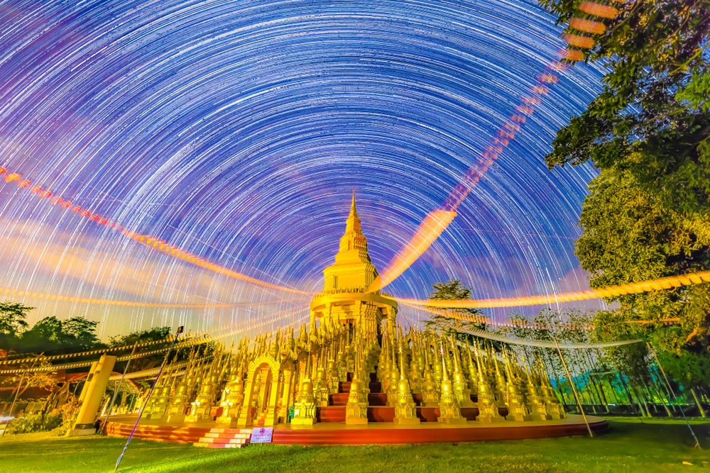 เจดีย์พุทธที่ล้อมรอบด้วยรูปปั้น ที่เที่ยวแก่งคอย ทองคำใต้เส้นทางดวงดาวที่มีชีวิตชีวาและท้องฟ้ายามค่ำคืนที่เต็มไปด้วยสีสันพร้อมต้นไม้ล้อมรอบฉาก ทิวทัศน์อันน่าหลงใหลที่เที่ยวชะอมแก่งคอย เหมาะสำหรับการผจญภัยครั้งถัดไปของคุณ.