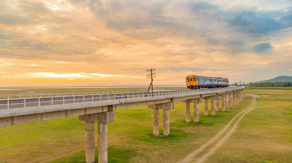 รถไฟเดินทางบนสะพานรถไฟคอนกรีตยกสูงเหนือที่ราบหญ้าในช่วงพระอาทิตย์ตกที่มีเมฆมาก นำเสนอ ที่เที่ยวแก่งคอย ทิวทัศน์ที่สวยงามเหมาะสำหรับทุกคนที่ต้องการสำรวจที่เที่ยวแก่งคอย.