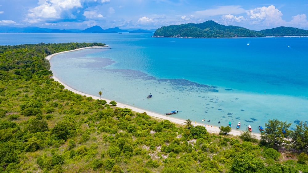 มุมมองทางอากาศของแนวชายฝั่งเขตร้อนในเที่ยวเกาะสมุย ที่มีความเขียวขจี หาดทราย น้ำทะเลสีฟ้าคราม เรือ ที่เที่ยวเกาะสมุย ทอดสมอ และเกาะห่างไกลภายใต้ท้องฟ้าที่มีเมฆบางส่วน.
