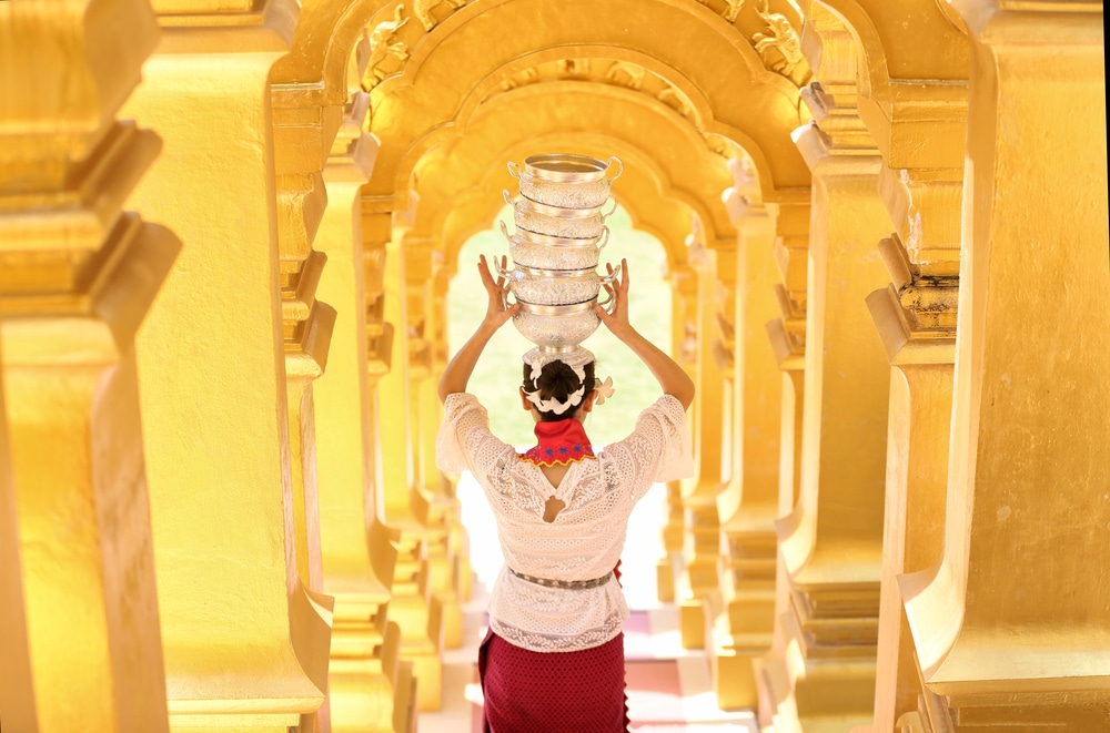 ผู้หญิงในชุดพื้นเมืองถือชามเงินซ้อนกันบนศีรษะขณะเดินผ่านซุ้มประตูสีเหลืองหรูหรา เผยให้เห็นเสน่ห์ของที่เที่ยว ที่เที่ยวแก่งคอย แก่งคอย.
