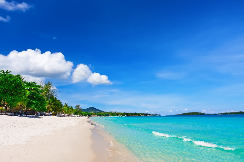 ชายหาดทรายที่มีน้ำทะเลสีฟ้าใส คลื่นเบาๆ และท้องฟ้าสีฟ้าใสที่มีเมฆปกคลุม ที่เที่ยวสมุย แห่งนี้ เรียงรายไปด้วยต้นไม้สีเขียวตลอดแนว ที่เที่ยวเกาะสมุย ชายหาดและมีเนินเขาสูงขึ้นไปเป็นฉากหลัง.